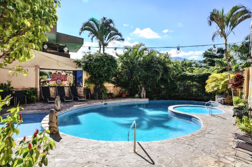 Sunny pool and Jacuzzi with waterfall mural, lounge chairs, and evening string lights.